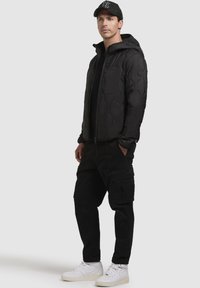 Schwarze Daunenjacke mit hexagonalem Steppmuster, Reißverschluss und Kapuze; kombiniert mit schwarzen Cargo-Hosen und weißen Turnschuhen.