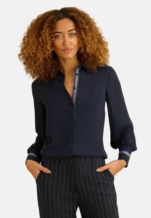 Nainen kiharat hiukset, yllään tummansininen pusero, jossa värikäs koriste, ja mustat pinstripe-housut, kädet taskuissa, yksiväristä taustaa vasten.