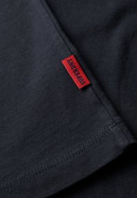 Camiseta de algodón de un oscuro color marino con una etiqueta bordada en rojo que dice "Superdry". Textura suave, cuello redondo estándar y mangas cortas.