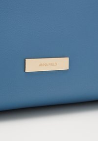 Sininen teksturoitu materiaali, jonka pinnalla on suorakulmainen kultaisen sävyinen tagi, johon on kaiverrettu "ANNA FIELD".