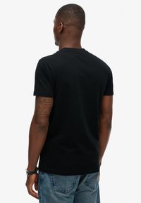 Sort bomulds T-shirt med korte ærmer, rund hals og et enkelt design. Bæret med blå denim jeans. Synlige tatoveringer på armen.