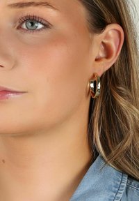 Guldörhängen med en polerad yta, med en slät, rund form. Bärs av en modell med långt hår, mot en neutral bakgrund.