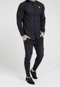 Camicia nera a manica lunga con chiusura a bottoni e colletto, abbinata a jeans neri strappati e sneaker in suede nere con logo dorato.