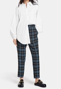 Witte lange mouwen blouse met een hoge kraag, gecombineerd met zwart-blauwe geblokte tapered broek en zwarte instap schoenen met gouden kettingdetail.