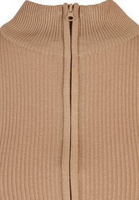 Gros plan sur un pull en maille côtelée beige avec un col montant et une fermeture éclair beige partiellement fermée au centre avant.