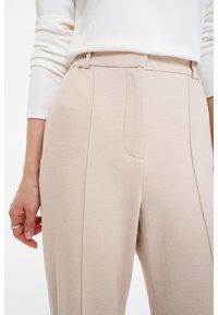 Helle beigefarbene, maßgeschneiderte Hose mit glatter Textur, ausgestattet mit einer einzigen Vordertasche, seitlichen Falten und einem elastischen Bund.