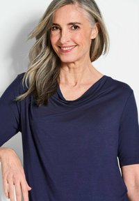 Navyblaues, drapiertes T-Shirt mit kurzen Ärmeln, glatter Textur und weitem Ausschnitt, getragen von einer lächelnden Person vor einem hellen Hintergrund.