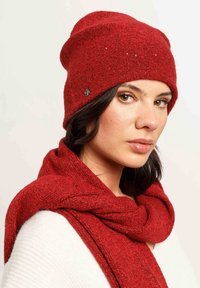 Red knit beanie with a textured finish and subtle embellishments, paired with a matching red scarf, shown against a light background.