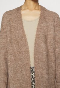 Brun stickad cardigan med en mjuk textur och öppet front. Bärs över en beige topp, ihop med mönstrade byxor med leopardtryck.