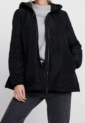 Femme portant une veste noire à capuche par-dessus un pull gris en maille et un jean gris foncé, debout avec une main dans la poche et une main le long du corps.