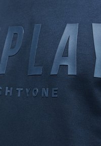 Ανυψωμένα σκούρα μπλε γράμματα που σχηματίζουν τη λέξη «PLAY» σε ύφασμα σκούρου μπλε χρώματος με μικρότερα μερικώς ορατά γράμματα από κάτω.