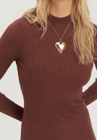 Femme blonde portant un haut à manches longues côtelé marron et un collier pendentif en forme de cœur en or sur une chaîne.
