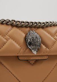 Sac en cuir tan avec un design matelassé, bandoulière en chaîne argentée et fermoir métallique orné en forme d'oiseau avec des détails décoratifs.