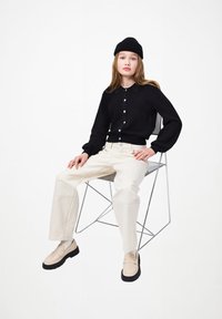 Joven con un gorro negro, suéter abotonado negro, pantalones crema y mocasines beige sentada en una silla de metal contra un fondo blanco.