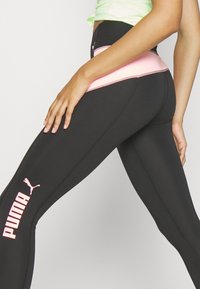 Černé sportovní legíny s růžovým pasem a bílým logem Puma. Materiál má hladkou, pružnou texturu s detaily švů.