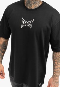Juodas medvilninis marškinėliai su baltu "TAPOUT" logotipu ant krūtinės. Trumpomis rankovėmis, apvaliu kaklu ir tekstūruotu audiniu.