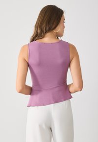 Woman wearing sleeveless lavender top and white pants, shown from the back against a plain light background.