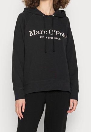 Personne portant un sweat à capuche noir Marc O’Polo avec cordons et texte blanc, debout devant un fond clair uni.