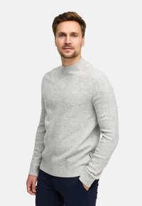 Uomo con capelli castano chiaro e barba, che indossa un maglione grigio chiaro e pantaloni scuri, in piedi con una mano in tasca su sfondo bianco.