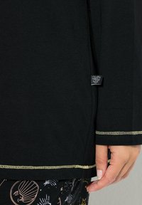 Černé tričko s dlouhými rukávy a zlatými švy, vyrobené z měkké bavlny. Na boku má štítek.