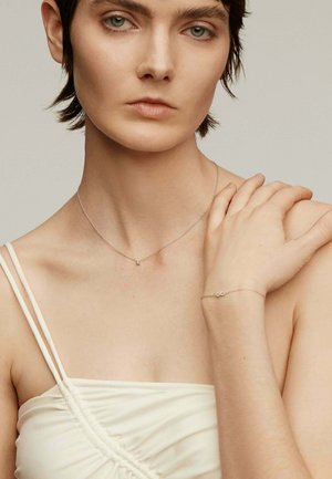 Collar de plata con un pequeño encanto, llevado en un cuello desnudo. También presenta una delicada pulsera de plata en una muñeca. Fondo claro y neutro.
