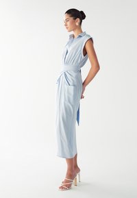 Hele sinine hõlmikolek, millel on krae, vööst rõhutatud ja küljetaskud, koos valgete rihmadega kontsaingadega. Sile tekstuur.