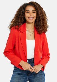 Blazer rosso con un design strutturato, chiusura a un bottone e rever a effetto. Tessuto morbido con maniche lunghe; indossato sopra una camicia bianca e jeans in denim.