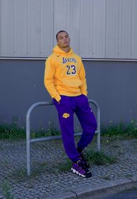 Moleton amarelo com o design "LAKERS 23", calças de treino roxas com o logótipo dos Lakers e ténis pretos com detalhes coloridos. Fundo com corrimão de aço interior.