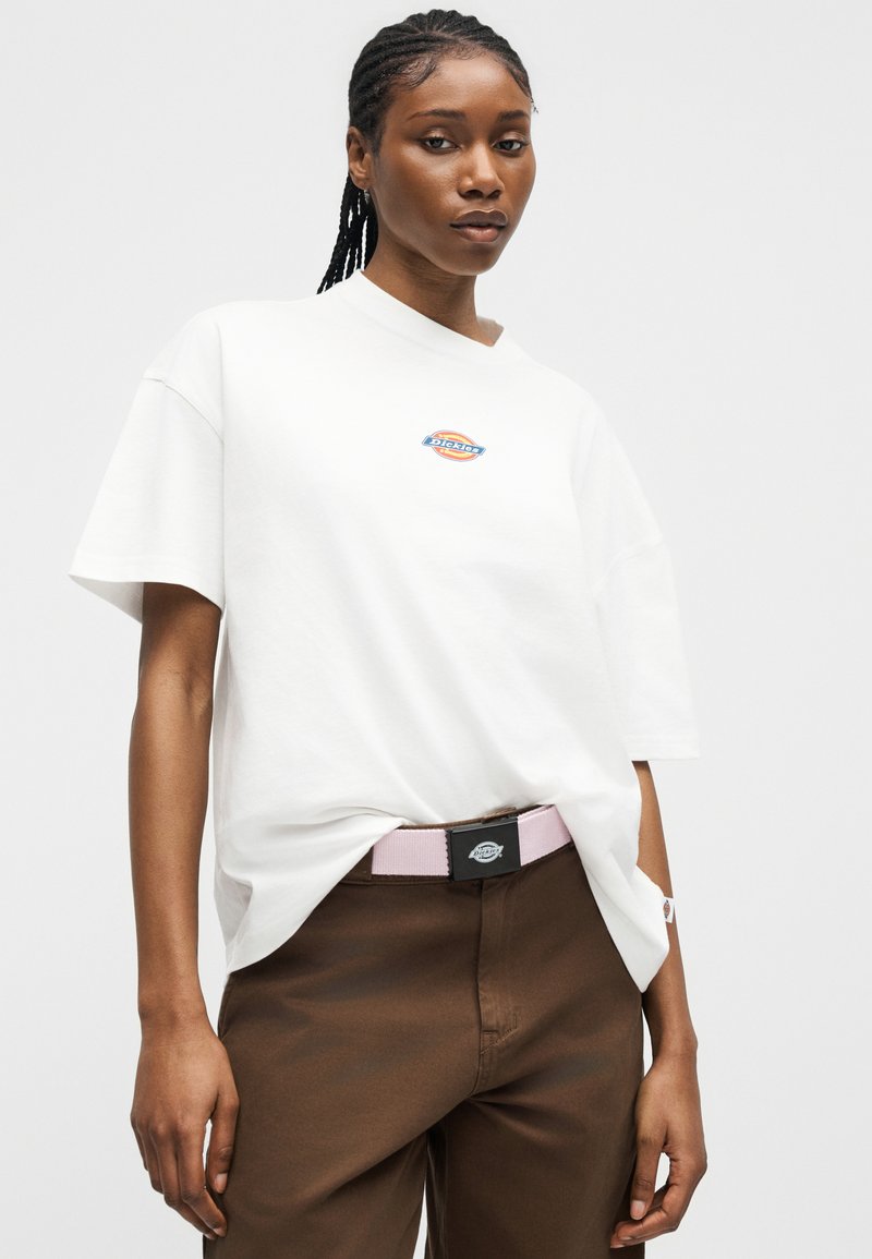 Donna con capelli intrecciati che indossa una maglietta bianca Dickies e pantaloni marroni con una cintura Dickies rosa chiaro, in piedi davanti a uno sfondo semplice.