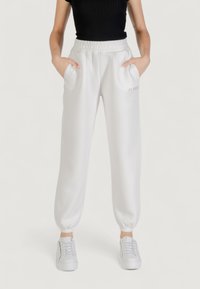 Pantaloni della tuta bianchi con una cintura a coste, polsini elastici, tasche laterali e un discreto marchio "BLESS" in testo chiaro. Abbinati a scarpe bianche.