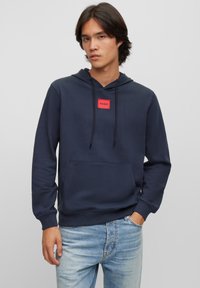 Mørkeblå hettegenser med et rødt rektangulært logopatch, snorhette, frontlomme, ribbede mansjetter og hem, laget av myk stoff.