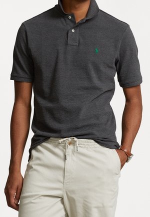 Graues Poloshirt mit strukturiertem Finish, Zwei-Knopf-Leiste und kleinem grünen Logo auf der linken Brust; kombiniert mit hellen Shorts.