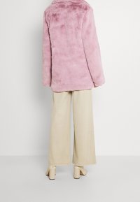 Rozige nepvachtjas met een ronde kraag, zachte textuur en oversized pasvorm, gecombineerd met brede beige broek en block-heeled schoenen.