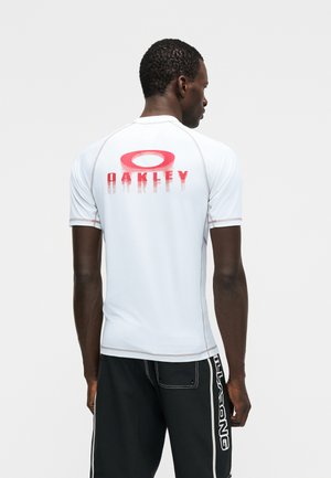 Ein Mann, der von hinten zu sehen ist, trägt ein weißes kurzärmliges Hemd mit rotem "Oakley"-Logo auf dem Rücken und schwarze Shorts mit weißem "Billabong"-Schriftzug.