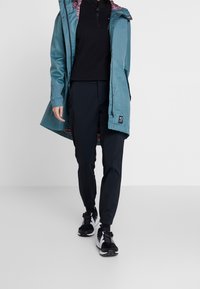 Light teal waterproof jacket with a patterned interior, black mid-layer, navy pants, and black athletic shoes with white accents.
