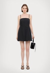 Abito nero senza maniche con corsetto aderente e gonna svasata. Il modello tiene una piccola borsa nera. Sandali neri con cinturini completano il look.