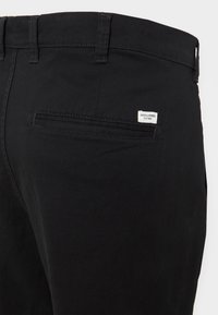 Schwarze Baumwollhose mit glatter Textur, ausgestattet mit einer Gesäßtasche mit Etikett und Gürtelschlaufen entlang der Taille.