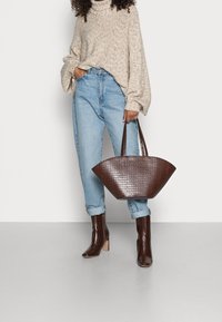 Braune Handtasche mit Krokodillederstruktur, breiter, geschwungener Form und doppelten Griffen, kombiniert mit hellblauen, hoch taillierten Jeans und einem beigen Pullover.