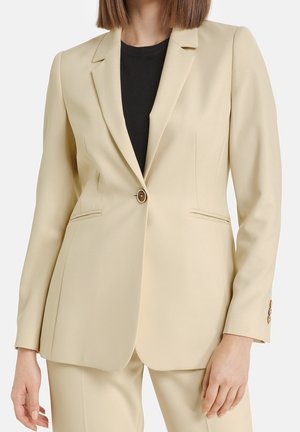 Femme portant un blazer beige cintré avec un bouton fermé sur un top noir, assorti à un pantalon beige.