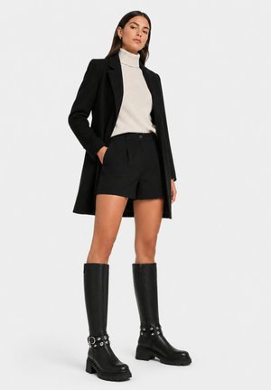 Kvinde iført sort blazer, sorte shorts, cremefarvet højhalset sweater og sorte knæhøje støvler med nitter, stående foran en ensfarvet baggrund.
