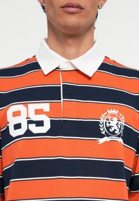 Polo riscado com cores laranja, azul marinho e branco. Possui um colarinho branco, um grande "85" bordado na parte da frente e um design de brasão.