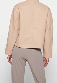 Personne portant une veste courte beige et un pantalon taupe, vue de dos sur un fond clair uni.