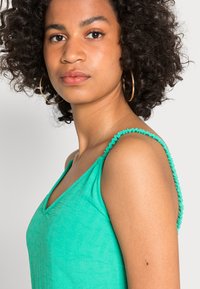 Top sem mangas em verde-água com decote em V e detalhes franzidos nas alças. Feito de um tecido texturizado e leve.