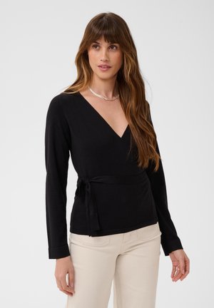 Femme aux longs cheveux bruns portant un haut noir à manches longues style cache-cœur et un pantalon beige clair sur un fond uni.