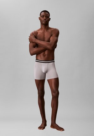 Uomo muscoloso in piedi a piedi nudi su sfondo grigio, indossa boxer grigio chiaro con fascia nera, braccia incrociate sul petto.