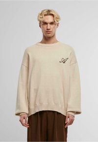 Pulover oversize bej din tricot cu manșete și tiv ondulate, având un logo brodat maro pe partea stângă din față. Textură netedă.