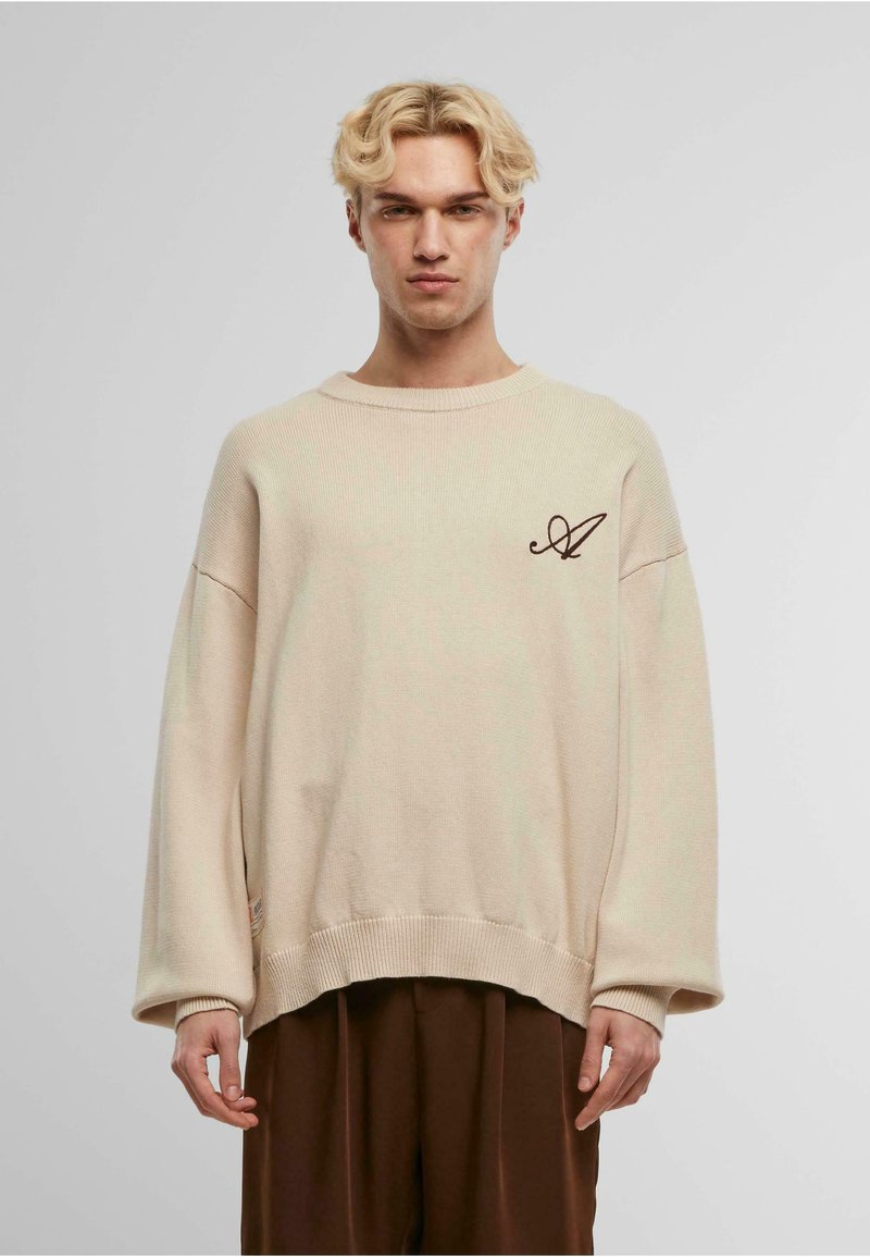 Beige oversized knit sweater with ribbed cuffs and hem, featuring a brown embroidered logo on the front left side. Smooth texture.