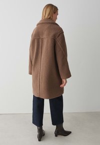 Manteau en polaire marron avec un large col, une coupe oversize et une légère texture. Le dos présente un détail de couture et est assorti à un jean foncé et des bottines.