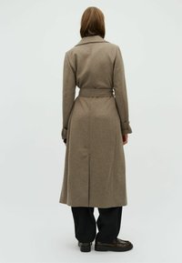 Lange bruine wollen jas met een ceintuur in de taille, gesneden kraag en zijsplitten. Gecombineerd met zwarte broeken en veterschoenen.