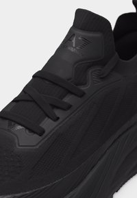 Baskets de sport noires en tissu texturé, lacets noirs et logo embossé sur la languette rembourrée, sur un fond clair uni.